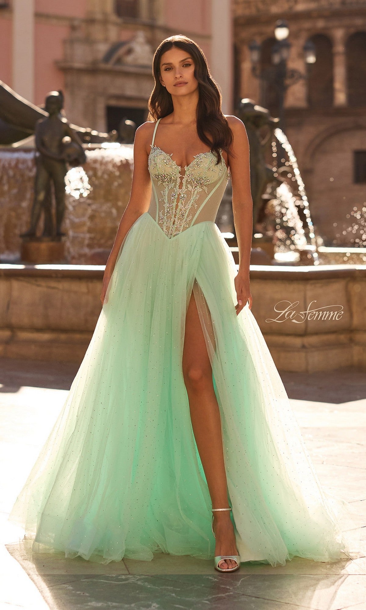 A woman stands outdoors by a fountain, wearing the La Femme 33860 long prom dress in light mint, featuring an embroidered corset bodice with thin straps, a high slit, and sparkling details.